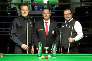 Jonathan Marwood and Rob Hall stand either side referee Stephen Harrison and in front of the trophies at the table.